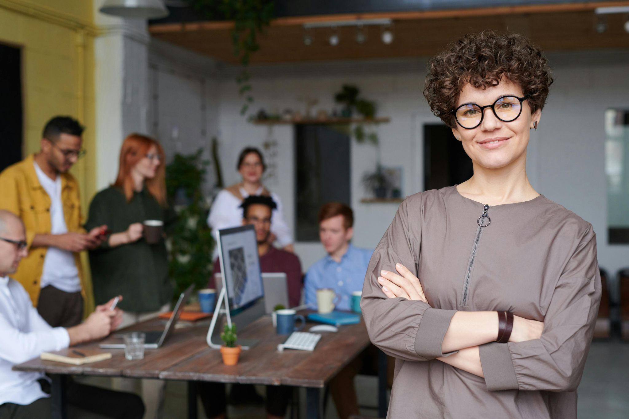 Confident businesswoman standing in a modern office with her team in the background, representing leadership in managing PEO benefits.