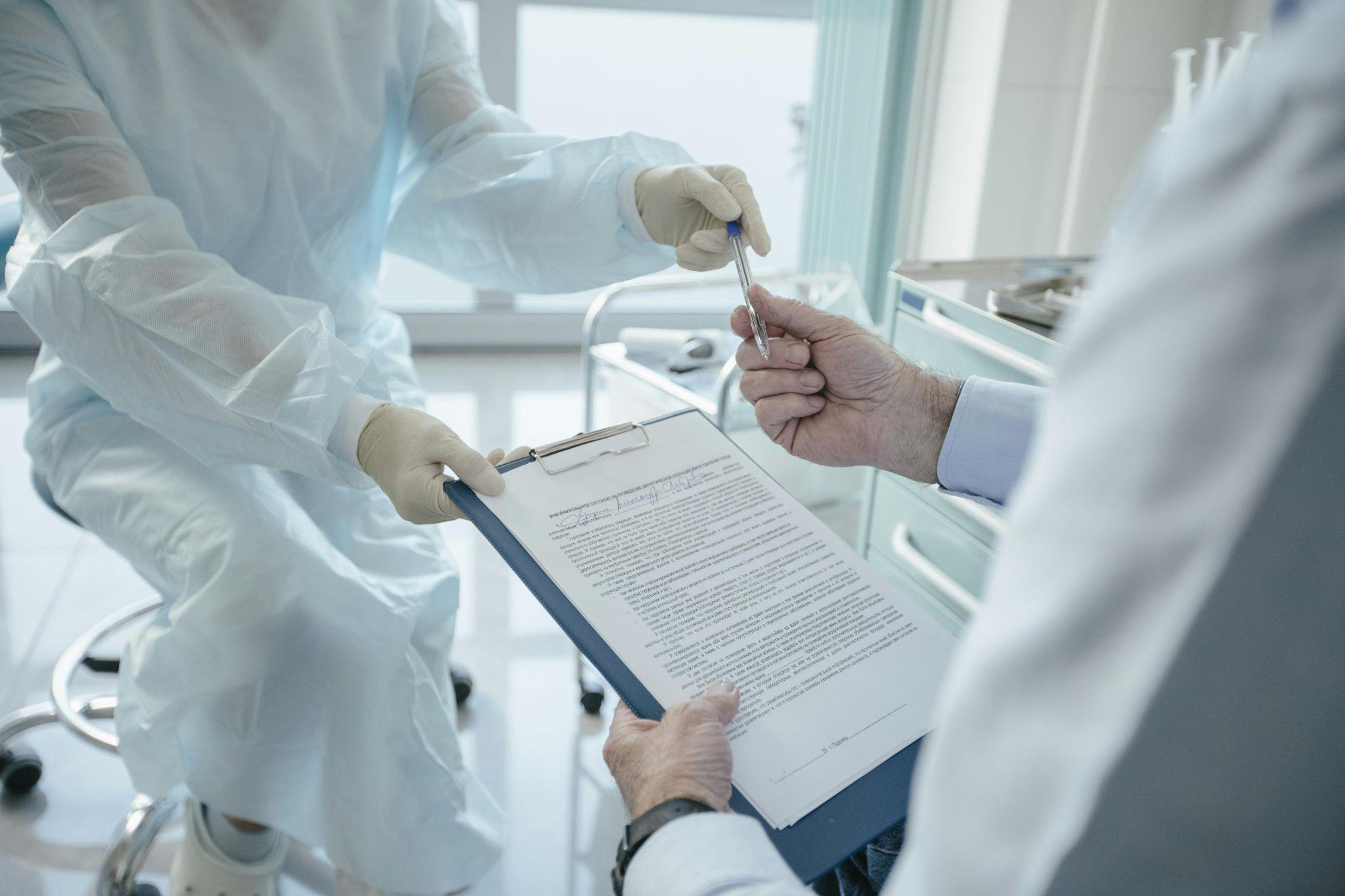 Doctor in protective clothing holding a clipboard while a patient hands over a pen, representing medical paperwork and healthcare processes