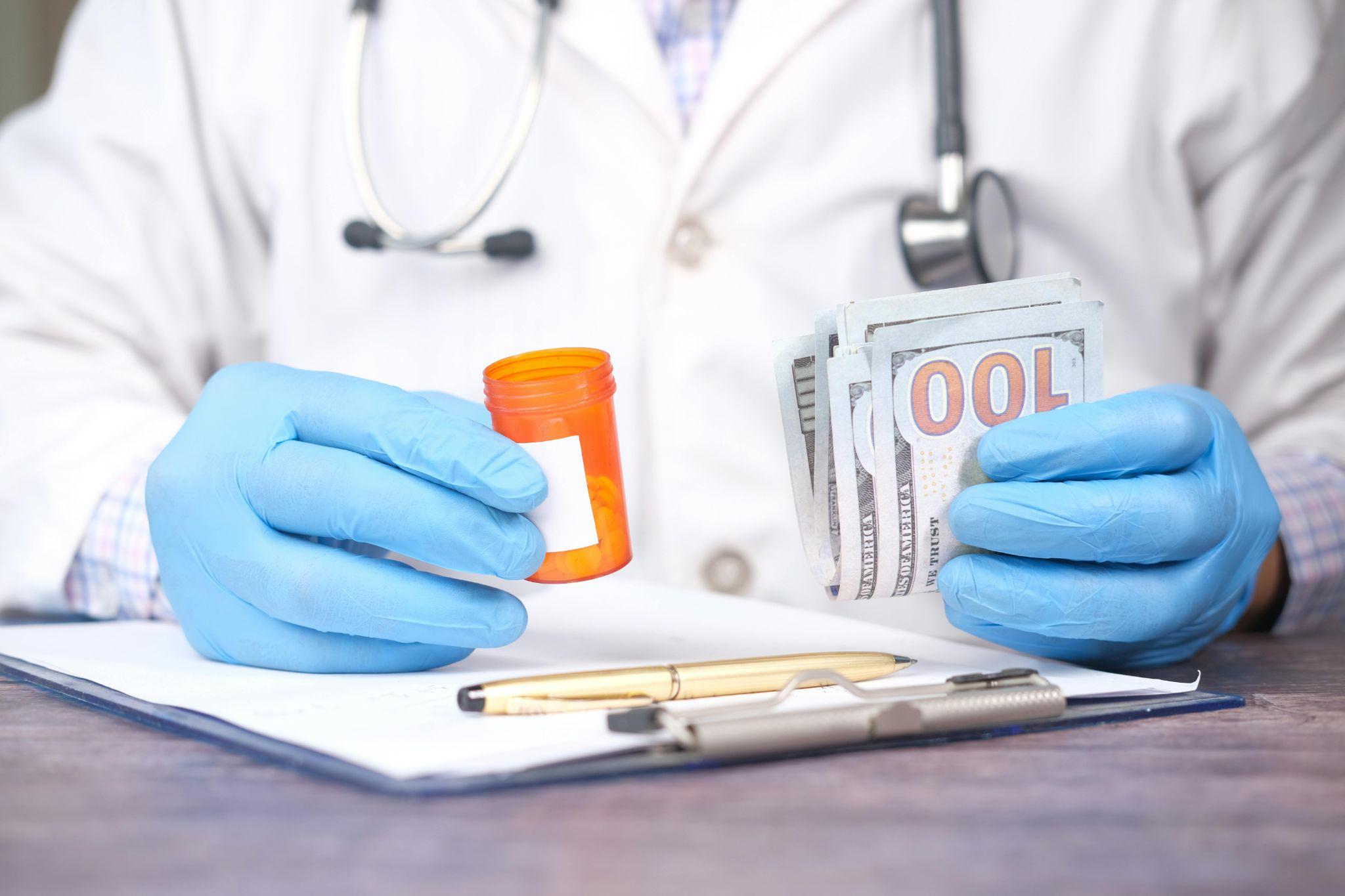 Doctor wearing blue gloves holding an orange prescription pill bottle in one hand and a stack of US hundred-dollar bills in the other, symbolizing the high cost of medication and healthcare expenses.