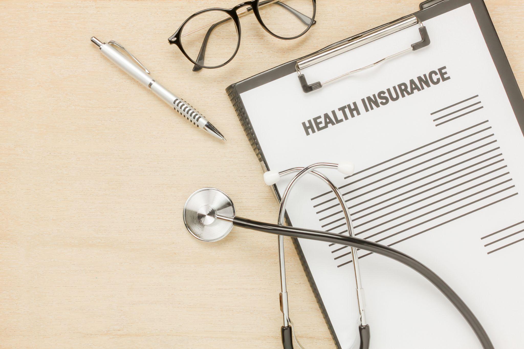 Clipboard with health insurance documents, glasses, and pen on a wooden desk, illustrating health insurance policy and paperwork