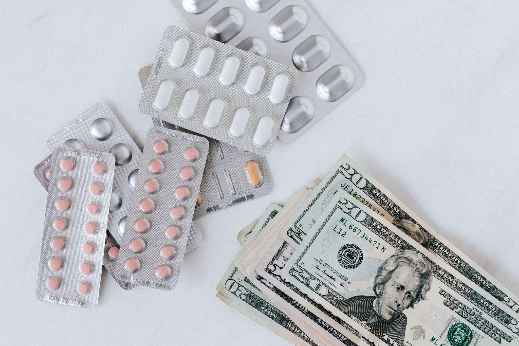Assorted pills in blister packs placed next to stacks of US dollar bills, representing the cost of healthcare and medical expenses.
